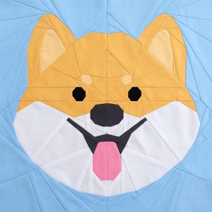 May include: A geometric quilt block featuring a Shiba Inu dog's face. The dog has a yellow and white face with black eyes and a black nose. The dog's tongue is pink. The background is light blue.