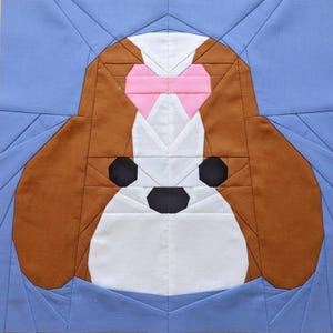Könnte beinhalten: Ein Stoffblock mit dem Gesicht eines Hundes, zusammengesetzt aus verschiedenen Stoffstücken. Der Hund hat braune Ohren, ein weißes Gesicht und eine rosa Schleife. Schwarze Formen bilden Augen und Nase, vor einem hellblauen Hintergrund.