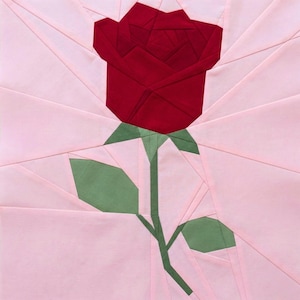 May include: A fabric art piece featuring a stylized red rose with green stem and leaves against a light pink background. The rose is the focal point, with detailed geometric petal construction. The background has a radiating pattern.