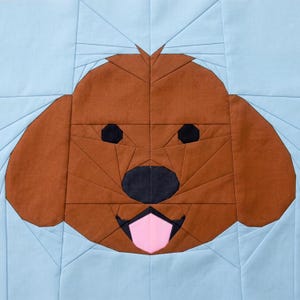 May include: A brown and black dog face quilt block with a light blue background. The dog's face is made of geometric shapes, with black eyes and nose, and a pink tongue. The quilt block is square and has a stitched pattern.