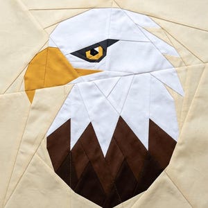 May include: A square fabric quilt block featuring a pieced design of a bald eagle head. The eagle has a white head, a sharp yellow beak, and a dark brown neck. The eye is black and yellow. The background is composed of light cream fabric pieces.