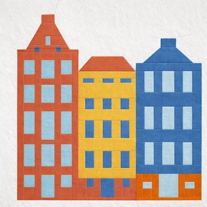 May include: A colorful fabric art piece featuring three stylized buildings. The buildings are in shades of orange, yellow, and blue, with light blue windows. The piece is likely a quilt block or a similar textile art project.