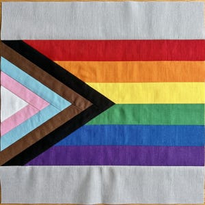 May include: A square fabric quilt block featuring the Progress Pride flag design. It includes horizontal stripes of red, orange, yellow, green, blue, and purple, with a chevron pattern on the left in black, brown, light blue, pink, and white.