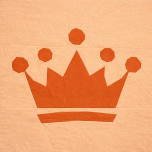 May include: A fabric crown design in orange, set against a light peach background. The crown features a series of points and circular accents, with a horizontal band at the base. The fabric shows visible seams. The image is a close-up.