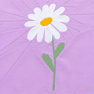 May include: A fabric art piece featuring a daisy with white petals and a yellow center. The flower has a green stem and leaves, set against a lavender background with geometric patterns. The artwork is a close-up view.
