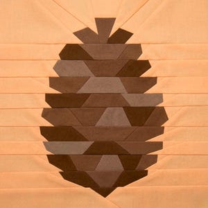 Könnte beinhalten: Ein geometrischer Quiltblock mit einem stilisierten Tannenzapfen-Design. Der Tannenzapfen besteht aus verschiedenen Brauntönen, vor einem hellpfirsichfarbenen Hintergrund. Das Design besteht aus zusammengenähten Stoffstücken, die einen texturierten Effekt erzeugen.