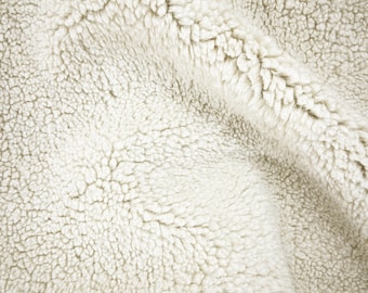 May include: Close-up of a cream-colored, textured fabric. The material has a dense, plush appearance with a slightly uneven surface. The fabric's texture is reminiscent of sheepskin or faux fur, with a soft, inviting look.