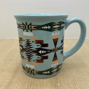 May include: A light blue ceramic mug with a handle, featuring a geometric pattern in shades of brown, white, and green. The mug has a cylindrical shape and is suitable for hot or cold beverages.
