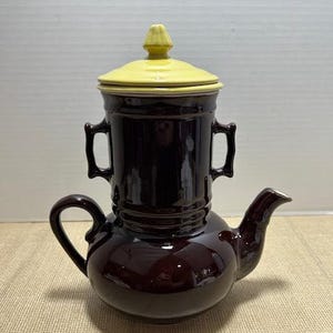 May include: A vintage brown ceramic teapot with a yellow lid. The teapot has a bulbous base, a curved spout, and a handle. The upper portion has two handles. The lid has a decorative finial.