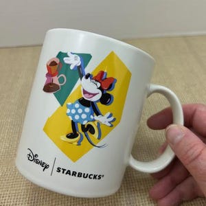 May include: White ceramic coffee mug featuring Minnie Mouse in a blue polka dot dress and a red bow. The mug has a yellow and teal background with the Disney and Starbucks logos.