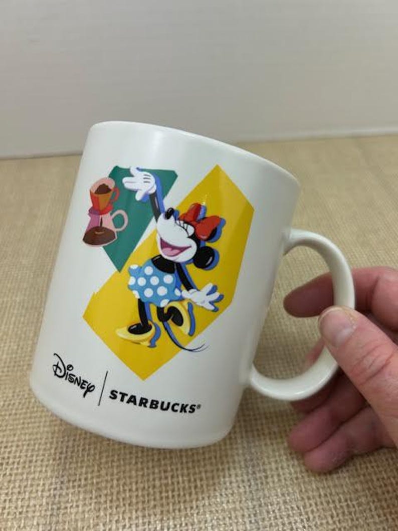 May include: White ceramic coffee mug featuring Minnie Mouse in a blue polka dot dress and a red bow. The mug has a yellow and teal background with the Disney and Starbucks logos.