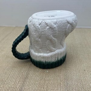 Pu&ograve; includere: Tazza in ceramica bianca con un design a maglia. La tazza ha un manico verde scuro e una fascia verde alla base. La tazza &egrave; progettata per assomigliare a un accogliente maglione invernale.
