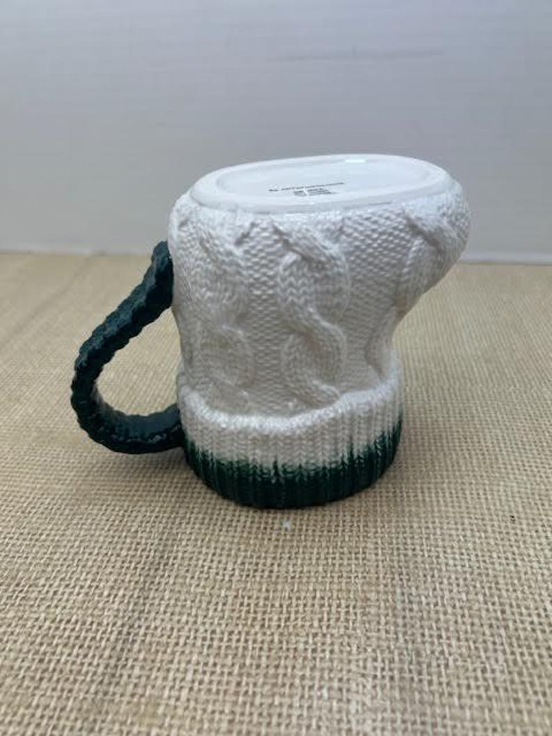 Pu&ograve; includere: Tazza in ceramica bianca con un design a maglia. La tazza ha un manico verde scuro e una fascia verde alla base. La tazza &egrave; progettata per assomigliare a un accogliente maglione invernale.