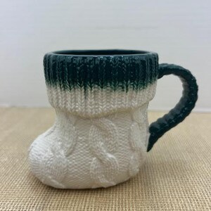 Pu&ograve; includere: Una tazza in ceramica a forma di calzino lavorato a maglia. Il bordo superiore e il manico sono verde scuro, che sfuma nel bianco con un motivo a trecce. La tazza &egrave; progettata per assomigliare a un caldo calzino invernale.