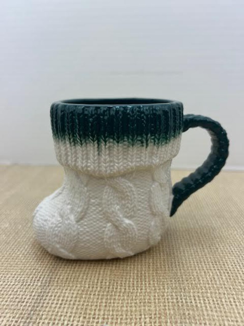 Pu&ograve; includere: Una tazza in ceramica a forma di calzino lavorato a maglia. Il bordo superiore e il manico sono verde scuro, che sfuma nel bianco con un motivo a trecce. La tazza &egrave; progettata per assomigliare a un caldo calzino invernale.