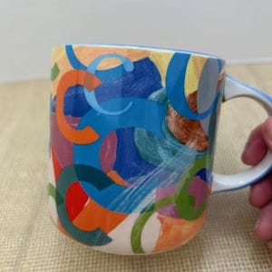May include: A ceramic coffee mug with a white interior and handle. The exterior features a colorful abstract design with orange, blue, green, and red shapes. The rim and handle are outlined in blue.