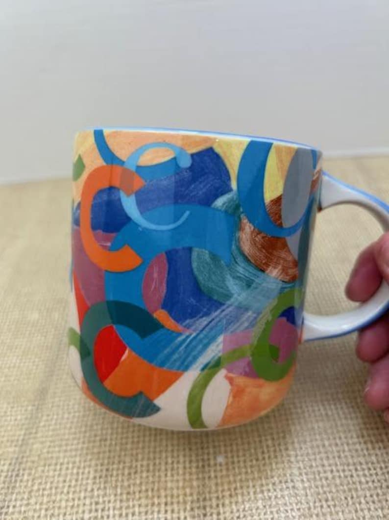 May include: A ceramic coffee mug with a white interior and handle. The exterior features a colorful abstract design with orange, blue, green, and red shapes. The rim and handle are outlined in blue.