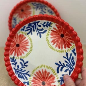 Pu&ograve; includere: Due piatti decorativi in ceramica con bordo smerlato rosso. I piatti presentano uno sfondo bianco con un motivo floreale rosso, blu e verde. I piatti sono progettati per servire cibo.