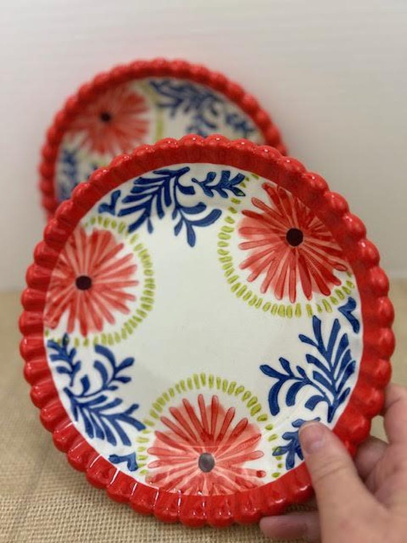 Pu&ograve; includere: Due piatti decorativi in ceramica con bordo smerlato rosso. I piatti presentano uno sfondo bianco con un motivo floreale rosso, blu e verde. I piatti sono progettati per servire cibo.