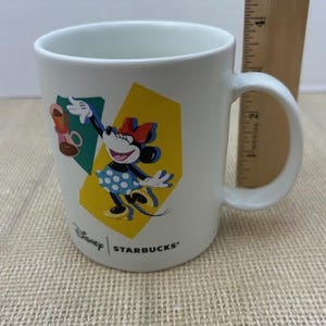 May include: White ceramic mug featuring Minnie Mouse in a polka dot dress, with a red bow and gloves. The mug has a yellow and green geometric design, with the Disney and Starbucks logos.