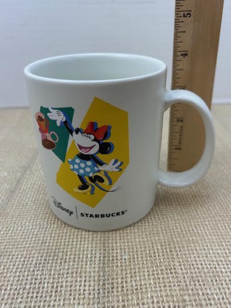 May include: White ceramic mug featuring Minnie Mouse in a polka dot dress, with a red bow and gloves. The mug has a yellow and green geometric design, with the Disney and Starbucks logos.