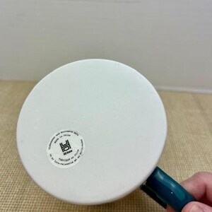 May include: White ceramic mug with a teal handle. The bottom of the mug has a circular sticker with text and a logo. The mug is dishwasher and microwave safe.