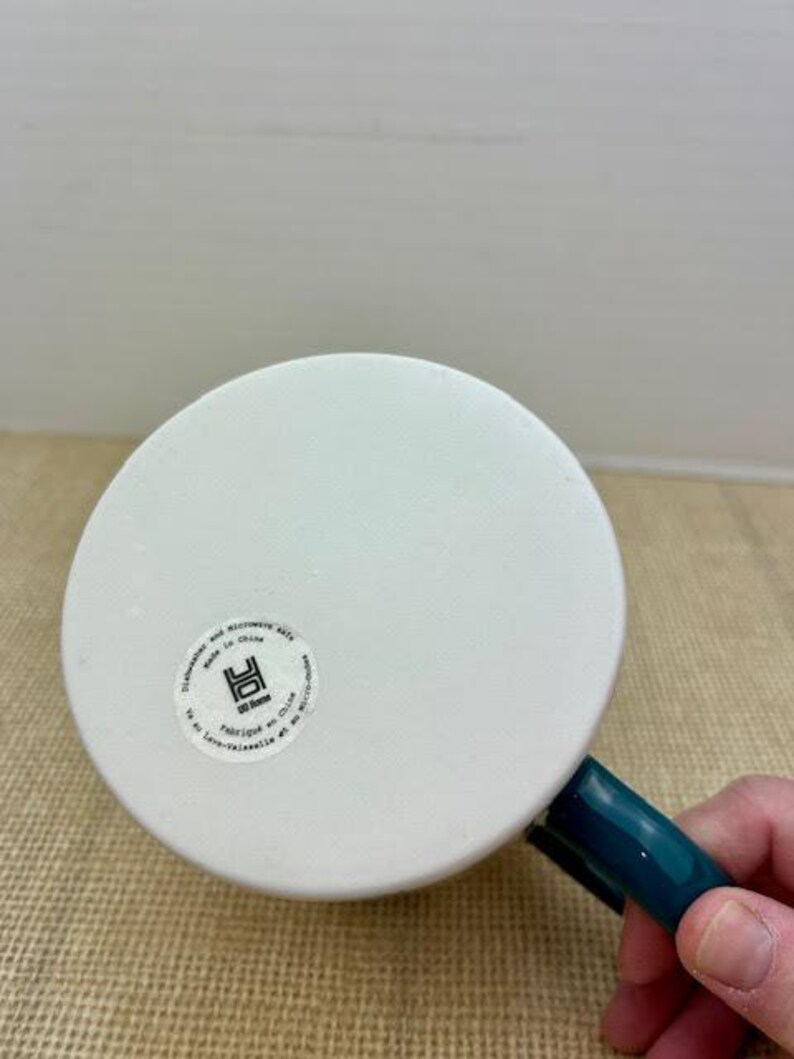 May include: White ceramic mug with a teal handle. The bottom of the mug has a circular sticker with text and a logo. The mug is dishwasher and microwave safe.