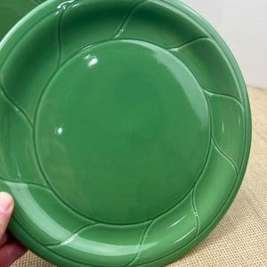 May include: A pair of glossy green ceramic plates. The plates have a central recessed area and a decorative leaf-like pattern around the rim. The plates are on a textured, neutral-colored surface.