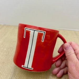May include: A red ceramic mug with a white letter "T" in the center. The mug has a curved handle and is set against a neutral background. The mug is a standard size for coffee or tea.
