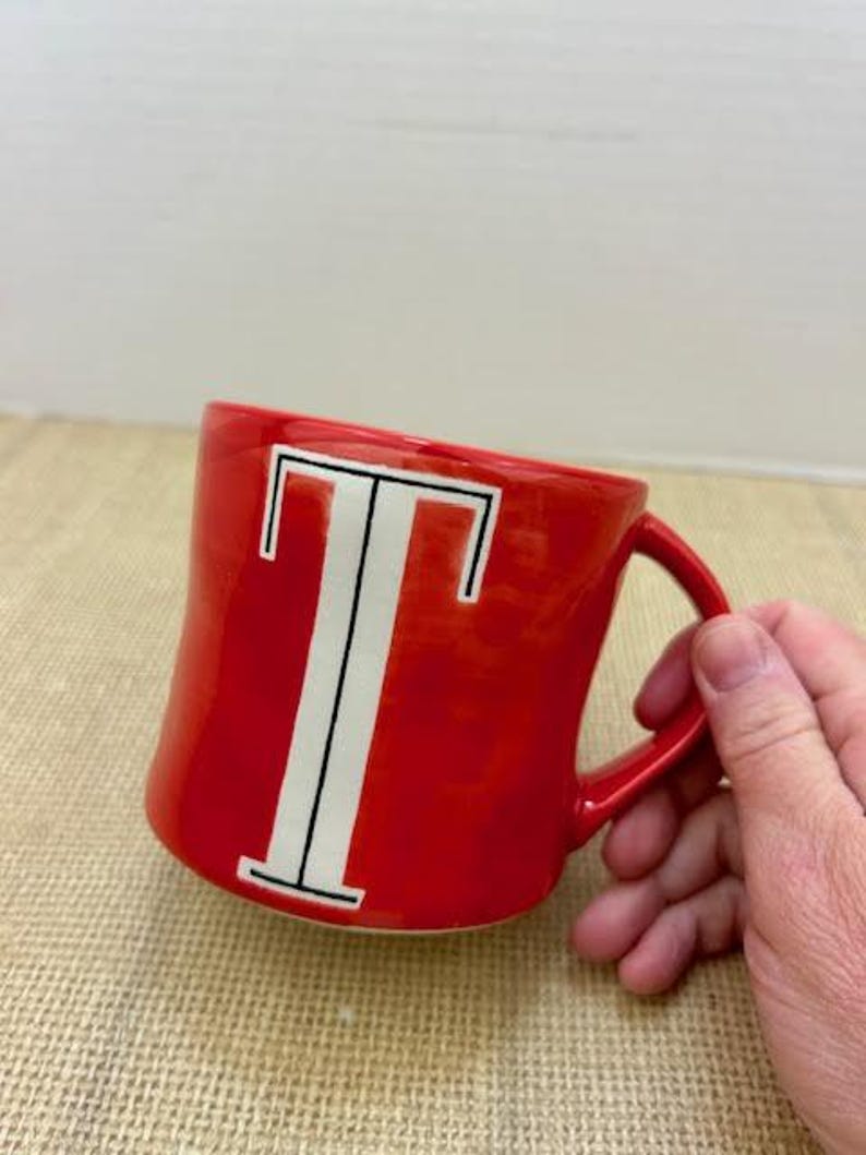May include: A red ceramic mug with a white letter "T" in the center. The mug has a curved handle and is set against a neutral background. The mug is a standard size for coffee or tea.
