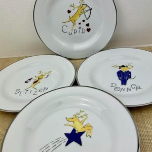 May include: Four white ceramic plates with thin silver rims, each featuring a unique cartoon reindeer illustration and name. The designs include Cupid with a bow, Blitzen holding a bottle, Donner in a blue suit, and Comet riding a blue star.