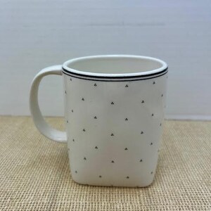 Puede incluir: Taza de cer&aacute;mica blanca con forma cuadrada y asa curva. La taza presenta una l&iacute;nea negra alrededor del borde y peque&ntilde;os patrones triangulares negros. La taza est&aacute; sobre una superficie beige.