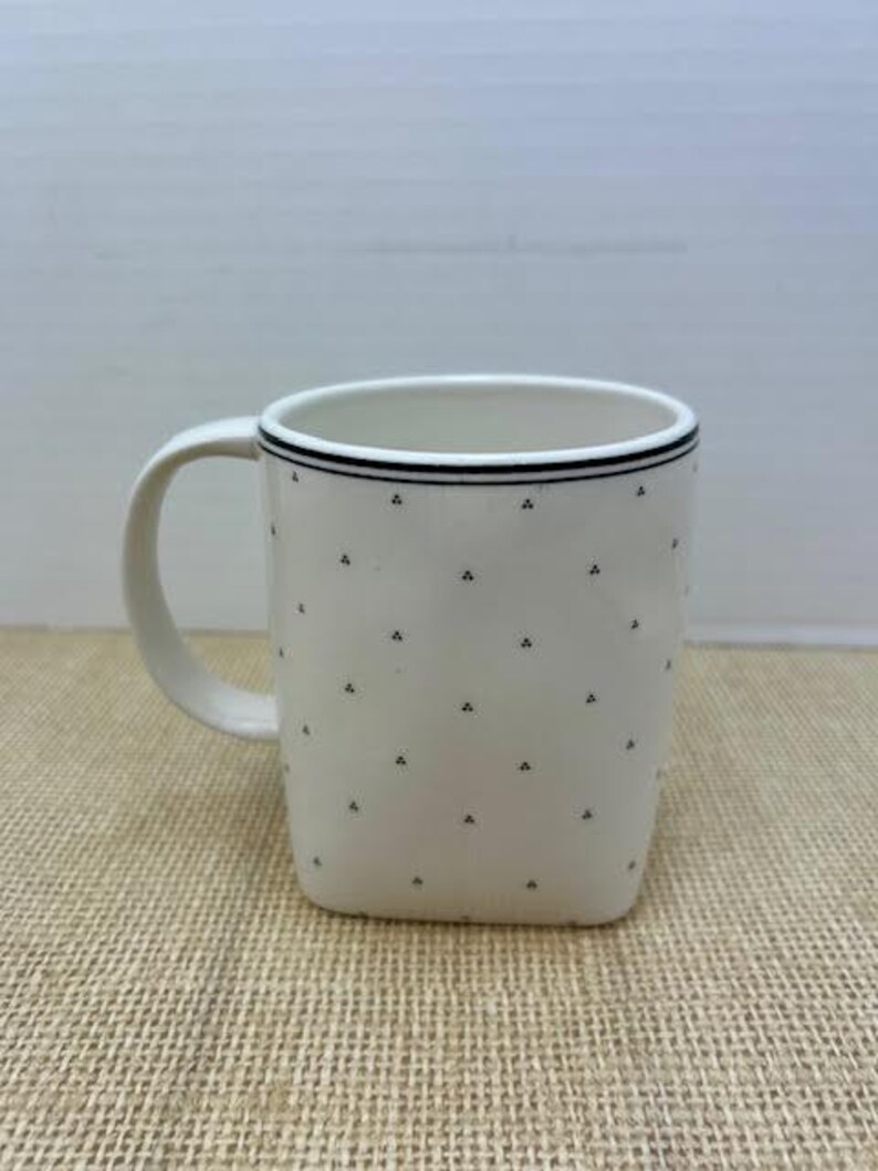 Puede incluir: Taza de cer&aacute;mica blanca con forma cuadrada y asa curva. La taza presenta una l&iacute;nea negra alrededor del borde y peque&ntilde;os patrones triangulares negros. La taza est&aacute; sobre una superficie beige.
