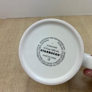 May include: White ceramic Starbucks mug. The base of the mug features the Starbucks logo, along with text indicating it is stoneware, microwave and dishwasher safe, and made in China. The mug holds 355 ml.