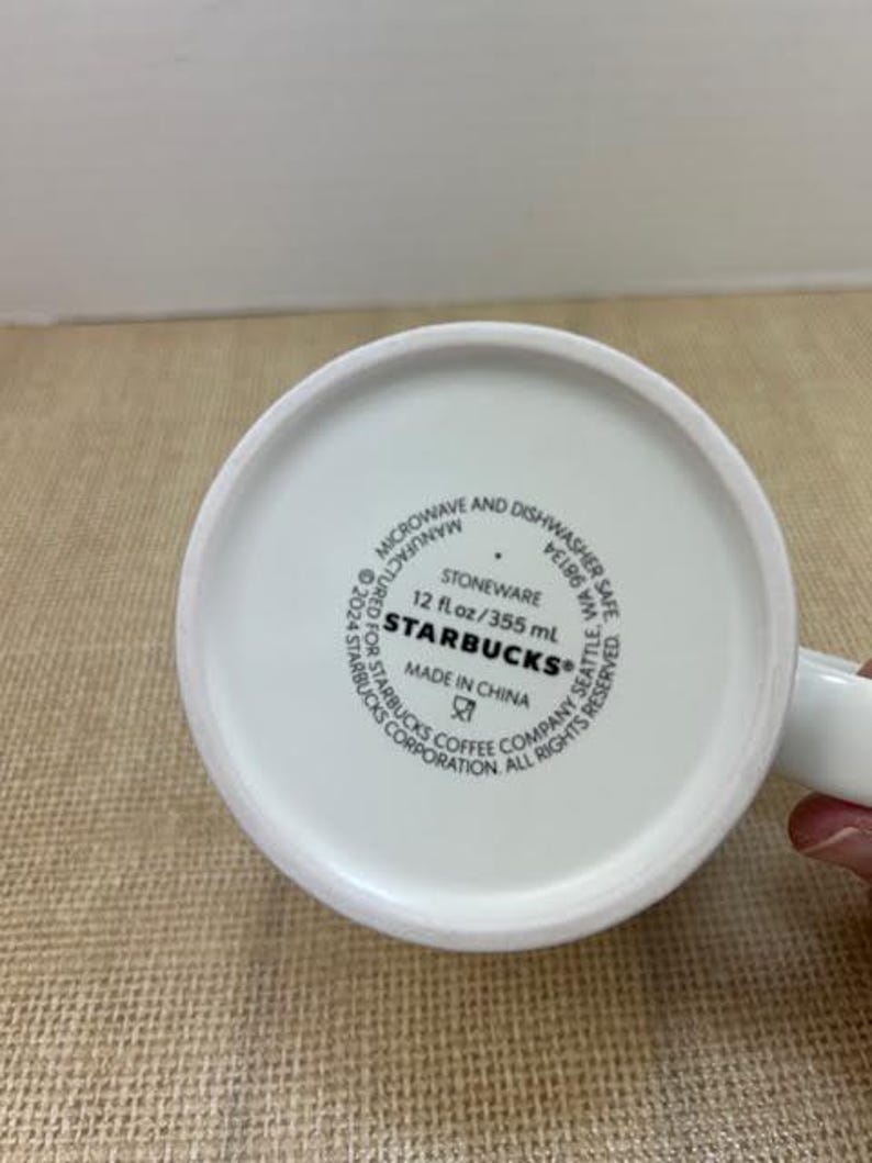 May include: White ceramic Starbucks mug. The base of the mug features the Starbucks logo, along with text indicating it is stoneware, microwave and dishwasher safe, and made in China. The mug holds 355 ml.