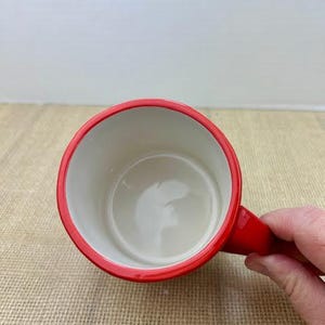 May include: A red coffee mug with a white interior. The mug has a red rim and handle. The mug is held by a person's hand.