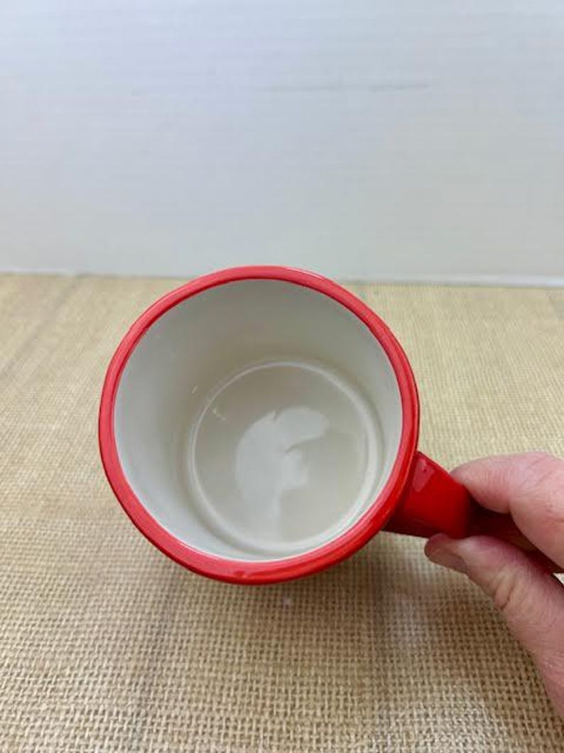 May include: A red coffee mug with a white interior. The mug has a red rim and handle. The mug is held by a person's hand.