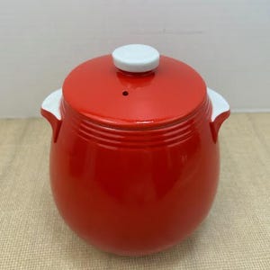 May include: A red ceramic jar with a lid and two white handles. The lid has a white knob and a small hole. The jar is a vibrant red color and has a glossy finish. The jar is likely for kitchen use.
