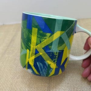 May include: A ceramic coffee mug with a white handle and a light green interior. The exterior features a vibrant abstract design with overlapping shapes in shades of blue, green, and yellow.