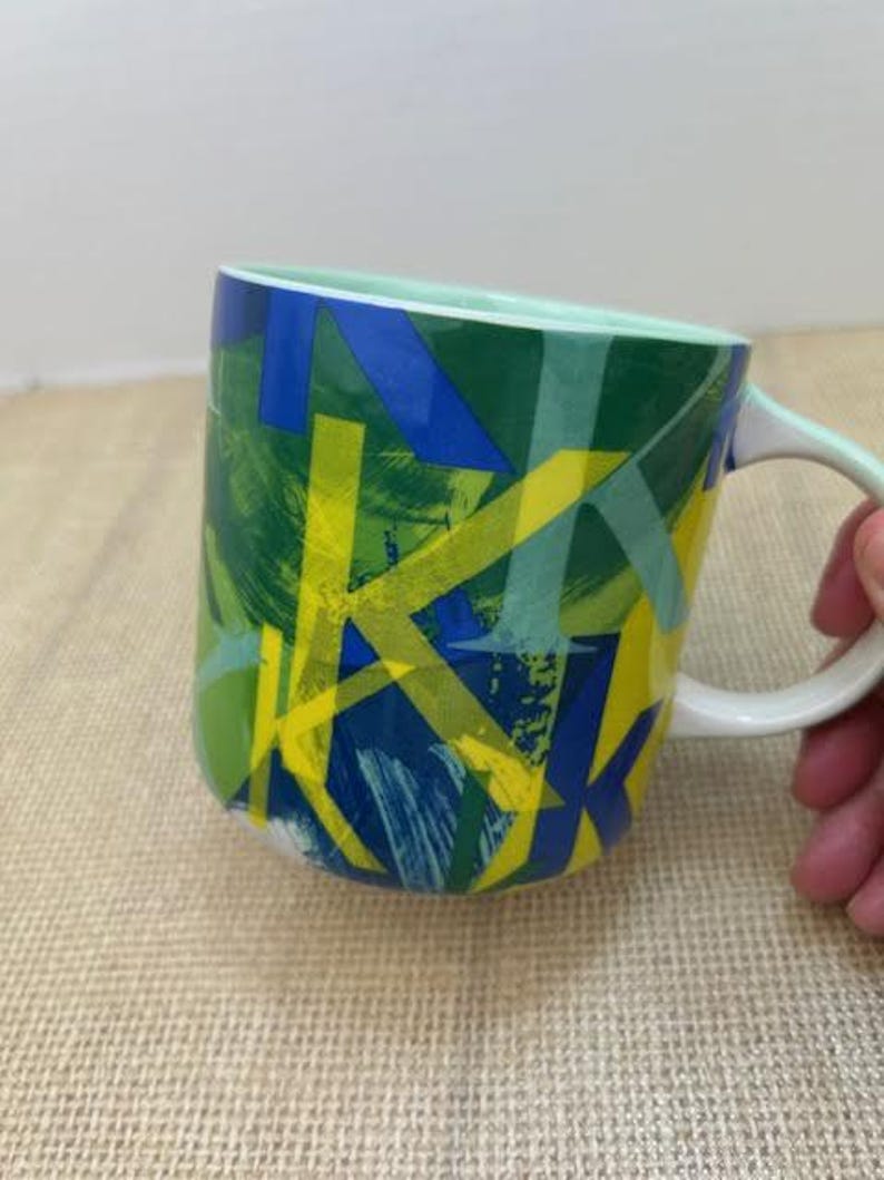 May include: A ceramic coffee mug with a white handle and a light green interior. The exterior features a vibrant abstract design with overlapping shapes in shades of blue, green, and yellow.