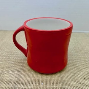 May include: A vibrant red ceramic mug with a white interior. The mug has a curved shape and a handle on the side. The mug is sitting on a textured surface.