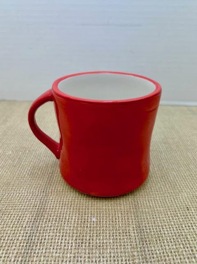 May include: A vibrant red ceramic mug with a white interior. The mug has a curved shape and a handle on the side. The mug is sitting on a textured surface.