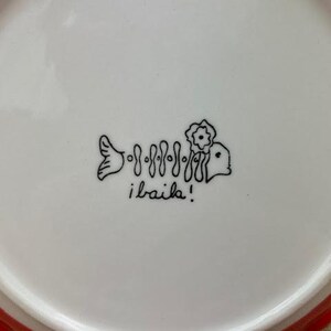 Pu&ograve; includere: Piatto in ceramica bianca con bordo smerlato rosso. Il piatto presenta un disegno a tratto nero di uno scheletro di pesce con un fiore e la scritta "i baila!"