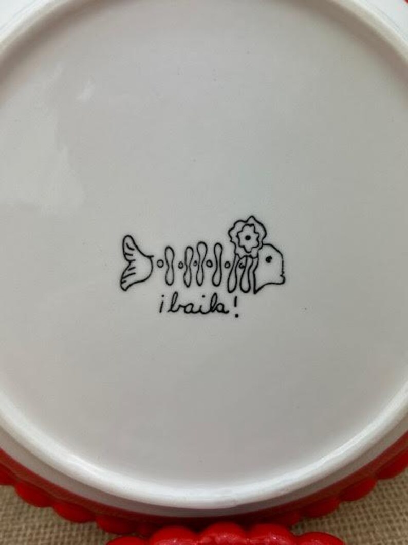 Pu&ograve; includere: Piatto in ceramica bianca con bordo smerlato rosso. Il piatto presenta un disegno a tratto nero di uno scheletro di pesce con un fiore e la scritta "i baila!"