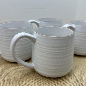May include: Four white ceramic mugs with a speckled finish and a ridged texture. Each mug has a large handle and a contrasting light brown rim. The mugs are arranged on a tan surface.