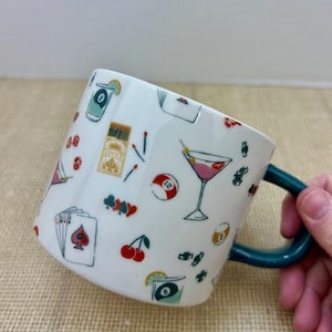 May include: White ceramic mug with a teal handle, decorated with colorful illustrations of cocktails, playing cards, cherries, and a pool ball. The mug has a whimsical design with a variety of small images.