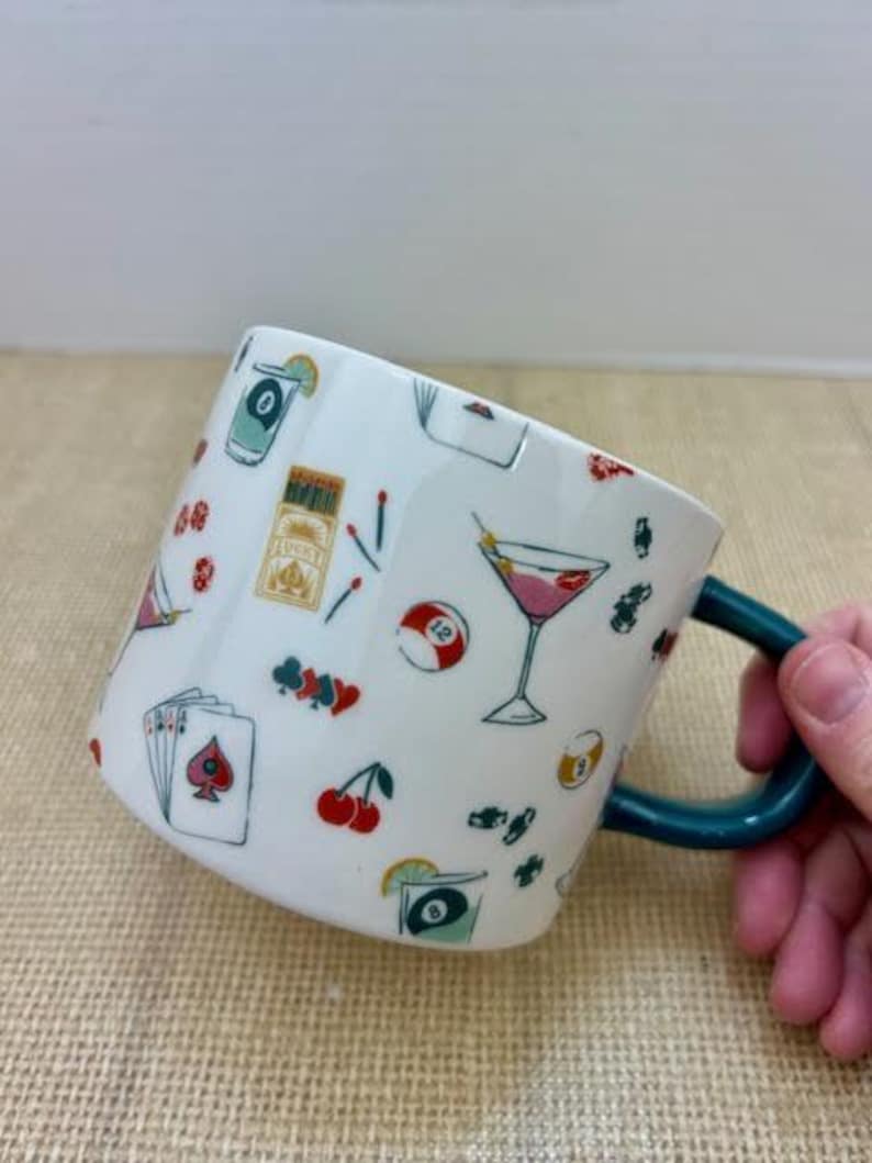May include: White ceramic mug with a teal handle, decorated with colorful illustrations of cocktails, playing cards, cherries, and a pool ball. The mug has a whimsical design with a variety of small images.