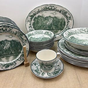 May include: A set of green and white patterned dinnerware with a woodland scene. The set includes a large dinner plate, a salad plate, a soup bowl, a teacup, and a saucer.