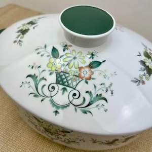 May include: A white porcelain tureen lid featuring a dark green circular handle and a floral basket motif. The design includes yellow, orange, and green flowers with intricate green scrollwork and foliage patterns painted on the curved surface.