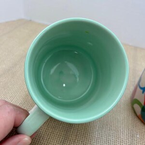 May include: A light green ceramic mug with a matching handle. The inside of the mug is also light green. The mug is sitting on a tan surface.